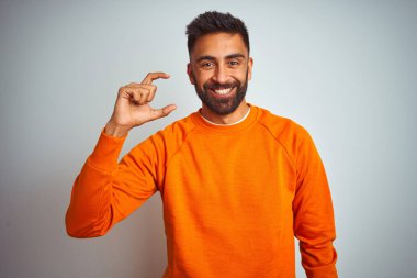 İzole edilmiş beyaz arka planda turuncu süveter giyen genç Hintli adam gülümsüyor ve kendinden emin bir şekilde el hareketi yapıyor parmaklarıyla işaret yapıyor ve kameraya bakıyor. Ölçü kavramı.