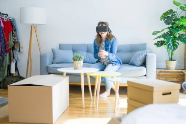 Yeni bir eve hareket karton kutular etrafında kanepede oturan bir simülasyon oyunu oynarken sanal gerçeklik gözlük giyen genç sarışın kadın
