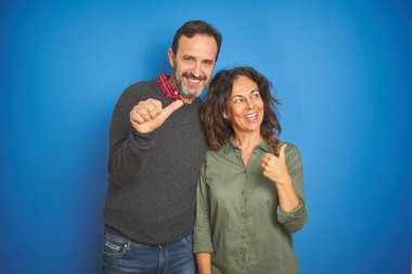 Güzel orta yaşlı çift, mavi arka planda yan yana durmuş gülümsüyor ve baş parmağıyla yan tarafa bakıyor..