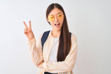 Gözlüklü ve sırt çantalı genç Çinli bir bayan izole edilmiş beyaz arka planda gülümsüyor ve kameraya gülümsüyor zafer işareti yapıyor. İki numara..