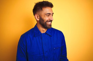 Sarı arka planda üniforma giyen yakışıklı Hintli işçi yüzünde gülümseme, doğal bir ifade ile yan yana bakıyor. Kendinden emin gülen.