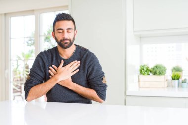 Evde rahat kazak giyen yakışıklı İspanyol adam yüzünde kapalı gözler ve minnettar jest ile göğsünde eller ile gülümsüyor. Sağlık konsepti.