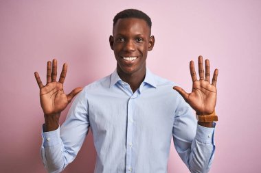 Mavi tişörtlü Afro-Amerikan bir adam pembe arka plan üzerinde duruyor ve parmaklarıyla on numarayı gösterirken kendinden emin ve mutlu gülümsüyor..