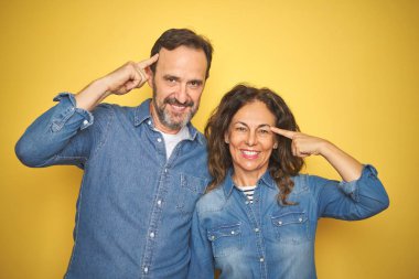 Güzel bir orta yaşlı çift, izole edilmiş sarı arka planda ayakta durup gülümsüyor, tek parmakla işaret ediyor, harika bir fikir ya da düşünce, iyi bir hafıza.