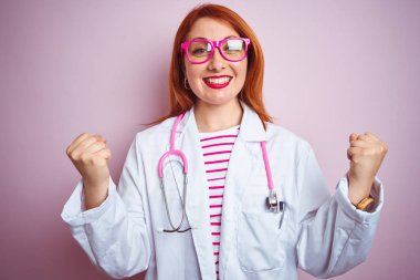 Genç kızıl saçlı kadın steteskop kullanıyor. Soyutlanmış pembe arka plan üzerinde duruyor. Kollarını kaldırıp, gözlerini açarak başarıyı kutluyor ve şaşırıyor. Kazanan konsept.