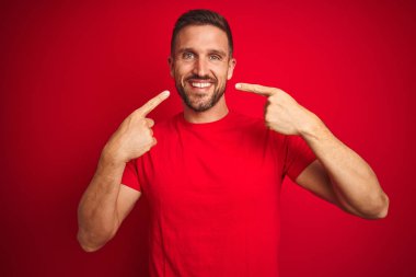 Kırmızı izole edilmiş arka plan üzerine gündelik tişört giyen genç yakışıklı adam gülümseyip, parmaklarıyla ve ağzıyla işaret ediyor. Diş sağlığı konsepti.