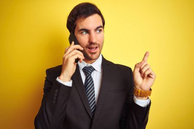 Akıllı telefondan konuşan genç yakışıklı iş adamı izole edilmiş sarı arka planda çok mutlu bir şekilde elini ve parmağını kenarı işaret ediyor.