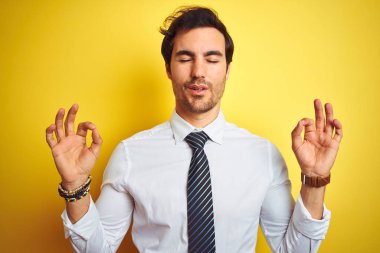 Zarif gömlek ve kravat giyen genç yakışıklı işadamı izole edilmiş sarı arka planda rahatla ve parmaklarıyla meditasyon yaparken gözleri kapalı gülümse. Yoga konsepti.