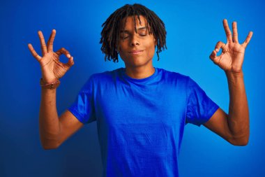Rastalı tişörtlü Afro-Amerikan bir adam izole edilmiş mavi arka planda dikiliyor rahatlıyor ve gözleri kapalı meditasyon yaparken parmaklarıyla gülümsüyor. Yoga konsepti.