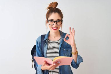 Sırt çantası takan kızıl saçlı kız izole edilmiş beyaz arka plan üzerine kitap okuyor tamam işareti yapıyor, mükemmel sembol.