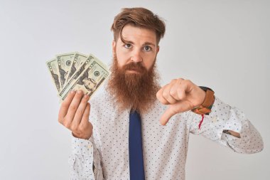Genç kızıl saçlı İrlandalı işadamı, izole edilmiş beyaz arka planda dolarlar tutuyor. Kızgın bir yüzü var. Negatif işaretler, başparmakları aşağıda, reddedilme kavramını gösteriyor.