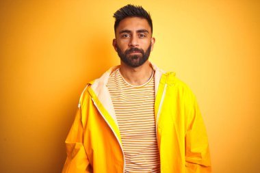 İzole edilmiş sarı arka planda duran yağmurluk giyen genç Hintli adam yüzünde ciddi bir ifadeyle rahatladı. Basit ve doğal bir şekilde kameraya bakıyorsun..