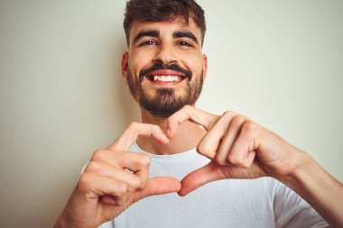 Dövmeli genç adam izole edilmiş beyaz arka plan üzerinde duruyor. Aşkla gülümsüyor. Elleriyle kalp sembolü ve şekli gösteriyor. Romantik konsept.