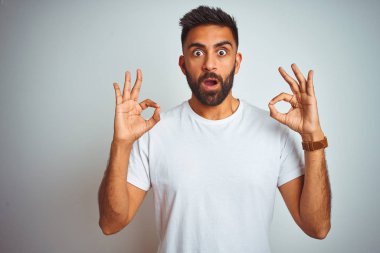 İzole edilmiş beyaz arka planda duran tişört giyen genç Hintli adam şaşırmış ve şaşırmış bir şekilde parmaklarıyla onay sembolü yapıyor. Çılgın ifade