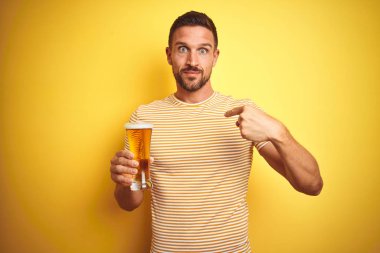 Genç yakışıklı bir adam, izole edilmiş sarı arka planda yarım litre bira içiyor.