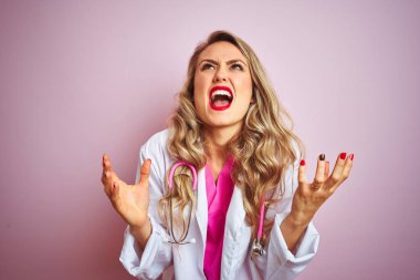 Pembe izole arka plan üzerinde steteskop kullanan genç ve güzel bir doktor. Deli gibi bağırıyor, bağırıyor, agresif bir ifade ve kollarla bağırıyor. Hayal kırıklığı konsepti.