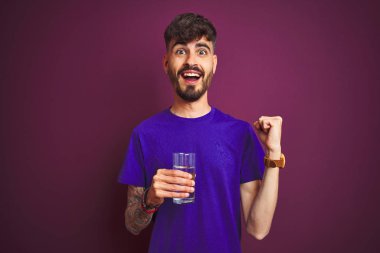 Dövmeli genç adam, izole edilmiş mor arka planda duran bir bardak su içerek gururla bağırıyor ve zaferi ve başarıyı kutluyor. Çok heyecanlı, coşkulu bir duygu.