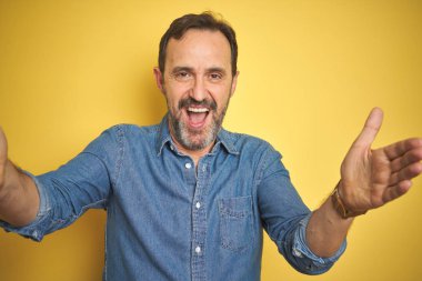 Sarı arka planda gri saçlı, yakışıklı, orta yaşlı bir adam kucaklaşmak için kollarını açmış kameraya bakıyor. Mutluluğu kucaklayan neşeli bir ifade..