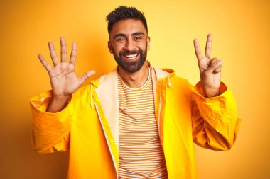 İzole edilmiş sarı arka planda duran yağmurluk giyen genç Hintli adam yedinci parmaklarını gösterirken kendinden emin ve mutlu bir şekilde gülümsüyor..