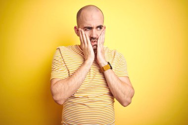 Sarı arka plan üzerine sıradan çizgili tişört giyen sakallı genç kel bir adam. Yüzü, depresyonu ve üzüntüyü örten yorgun ve tedirgin eller.