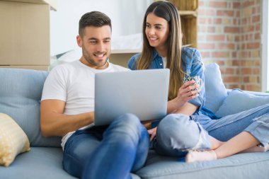 Genç çift yeni bir ev bilgisayar laptop kullanarak kanepede oturan rahatlatıcı, yeni daireye taşımak için mutlu gülümseyen hareket