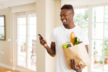Bakkaliye tam bir kağıt torba tutarak ve online satın alma smarpthone kullanarak Afrika adam gülümseyerek app kullanarak