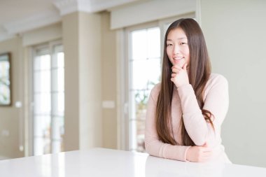Güzel Asyalı kadın emin çapraz kollar ve çeneme kaldırdı el ile gülümseme ile kameraya bakarak beyaz masada rahat kazak giyiyor. Pozitif düşünce.