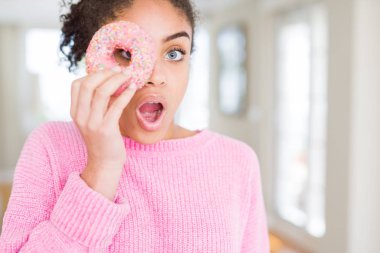 Genç Afro-Amerikalı kız pembe donut yiyor. Şaşırmış bir suratla korkmuş ve korku ifadesinden heyecanlanmış.