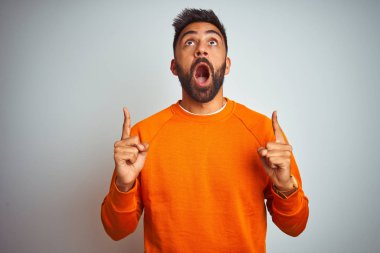 İzole edilmiş beyaz arka plan üzerinde turuncu süveter giyen genç Hintli adam şaşırmış ve şaşırmış bir şekilde yukarı baktı ve parmaklarıyla işaret ederek kollarını kaldırdı..