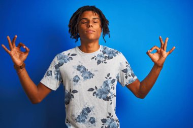 Rasta desenli tişörtlü Afro-Amerikan bir adam izole edilmiş mavi arka plan üzerinde rahatla ve gözleri kapalı meditasyon yaparken parmaklarıyla gülümse. Yoga konsepti.