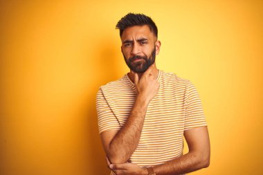 İzole edilmiş sarı arka planda duran tişört giyen genç Hintli adam kameraya bakarken kollarını kavuşturup ellerini çenesine kaldırarak gülümsüyor. Olumlu düşünüyorum..