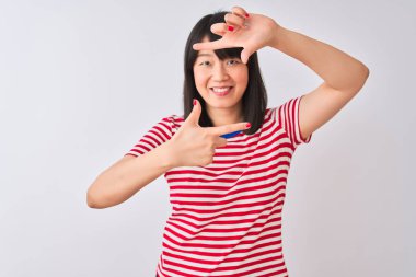 İzole edilmiş beyaz arka plan üzerinde kırmızı çizgili tişört giyen genç, güzel Çinli kadın gülümsüyor ve mutlu yüzlü elleri ve parmakları olan bir çerçeve yapıyor. Yaratıcılık ve fotoğrafçılık kavramı.