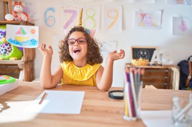Gözlüklü ve tek boynuzlu at diademi takmış güzel bir çocuk anaokulunda oturmuş kollarını kaldırmış ve gözleri kapalı heyecanla haykırarak başarının çılgınlığını kutluyor. Kazanan konsept