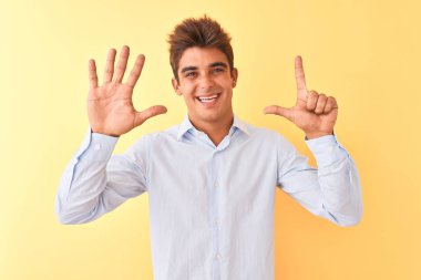 Genç yakışıklı iş adamı zarif bir gömlek giyerek izole edilmiş sarı arka plan üzerinde 7 numaralı parmaklarıyla işaret ederken kendinden emin ve mutlu bir şekilde gülümsüyor..