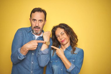 Güzel orta yaşlı çift, izole edilmiş sarı arka planda dikiliyorlar. Aceleyle saati, sabırsızlığı izlemek ve rahat bir ifadeyle kameraya bakmak istiyorlar.