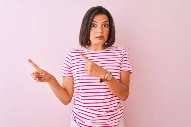 İzole edilmiş pembe arka plan üzerinde duran çizgili tişört giyen genç ve güzel bir kadın iki eliyle endişeli ve endişeli bir ifadeye işaret ediyor.