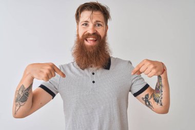 Genç, kızıl saçlı, gri polo giyen, izole edilmiş beyaz arka planda duran, kendine güvenen, gülümseyen, kendini gururlu ve mutlu gösteren bir adam..