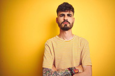 İzole edilmiş sarı arka plan üzerinde duran çizgili tişörtlü genç adam şüpheci ve gergin, yüzünde çapraz kollu tasvip etmeyen ifade. Negatif kişi.