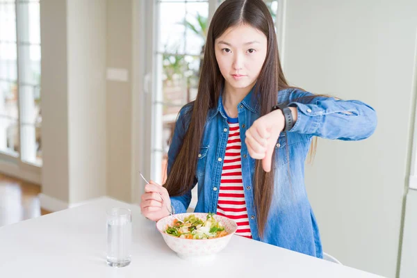 Beautiful Asian woman eating healthy pasta salad with angry face, negative sign showing dislike with thumbs down, rejection concept