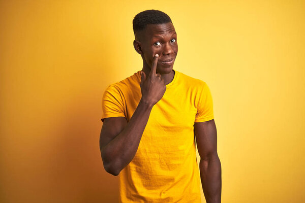 Young african american man wearing casual t-shirt standing over isolated yellow background Pointing to the eye watching you gesture, suspicious expression