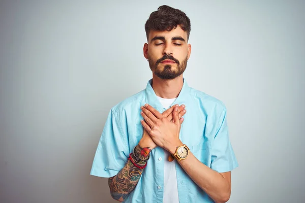 Young man with tattoo wearing blue shirt standing over isolated white background smiling with hands on chest with closed eyes and grateful gesture on face. Health concept.