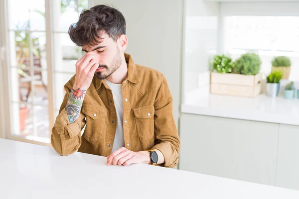 Young man wearing casual jacket sitting on white table tired rubbing nose and eyes feeling fatigue and headache. Stress and frustration concept.