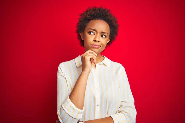 Young beautiful african american woman with afro hair over isolated red background with hand on chin thinking about question, pensive expression. Smiling with thoughtful face. Doubt concept.