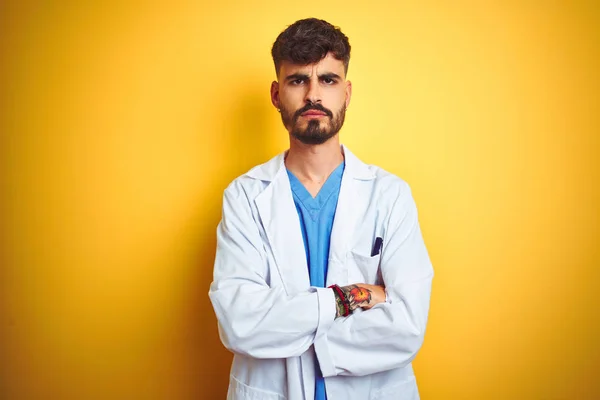 Young doctor man with tattoo standing over isolated yellow background skeptic and nervous, disapproving expression on face with crossed arms. Negative person.