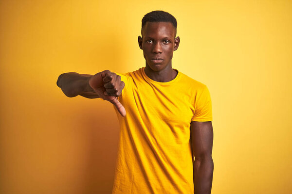 Young african american man wearing casual t-shirt standing over isolated yellow background looking unhappy and angry showing rejection and negative with thumbs down gesture. Bad expression.