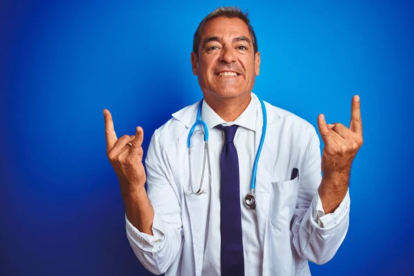 Handsome middle age doctor man wearing stethoscope over isolated blue background shouting with crazy expression doing rock symbol with hands up. Music star. Heavy concept.