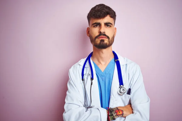 Young doctor man with tattoo wearing stethocope standing over isolated pink background skeptic and nervous, disapproving expression on face with crossed arms. Negative person.