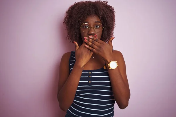 孤立したピンクの背景に縞模様のTシャツの眼鏡をかけている若いアフリカのアフロ女性は間違いのために手で口をカバーショックを受けた 秘密概念 — ストック写真