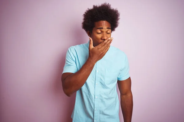 Young american man with afro hair wearing blue shirt standing over isolated pink background bored yawning tired covering mouth with hand. Restless and sleepiness.