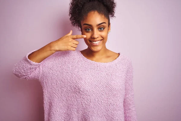 Young african american woman wearing winter sweater standing over isolated pink background Pointing with hand finger to face and nose, smiling cheerful. Beauty concept